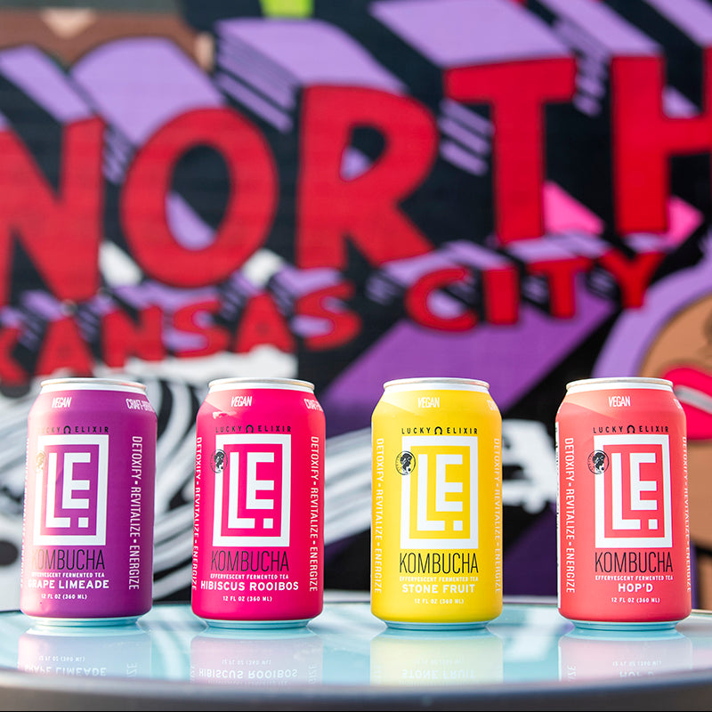 Four cans of Lucky Elvis kombucha on a reflective surface with a colorful 'North Kansas City' sign in the background.