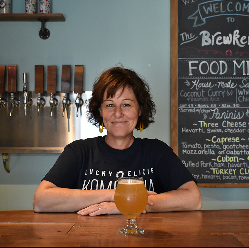 Woman sitting at a bar with a glass of kombucha, in front of a chalkboard menu.