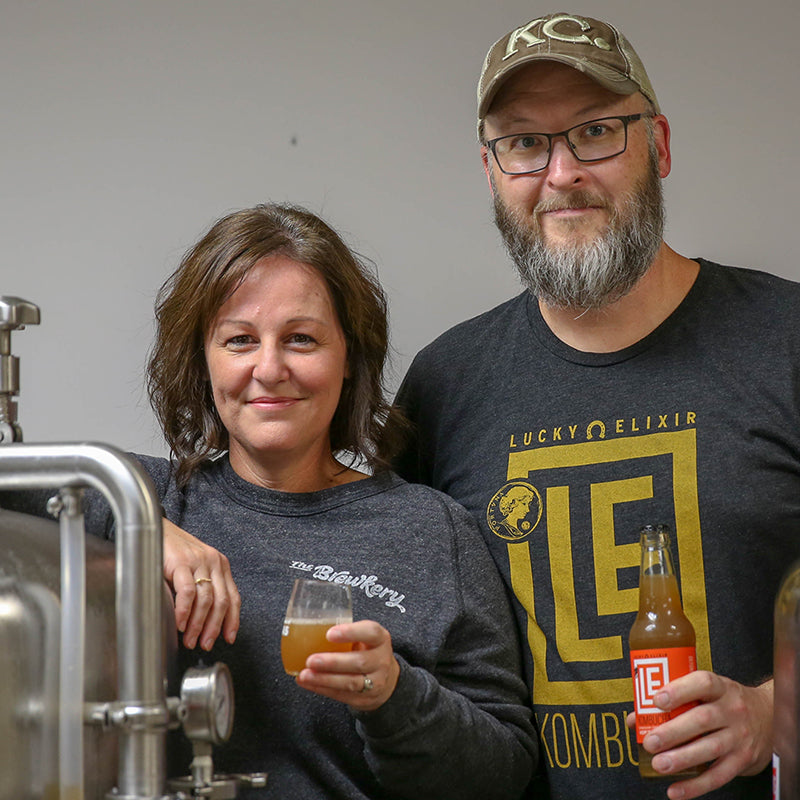Two people, a man and a woman, standing next to each other holding a glass of kombucha and a bottle. The man is wearing a 'Lucky Elixir' t-shirt.