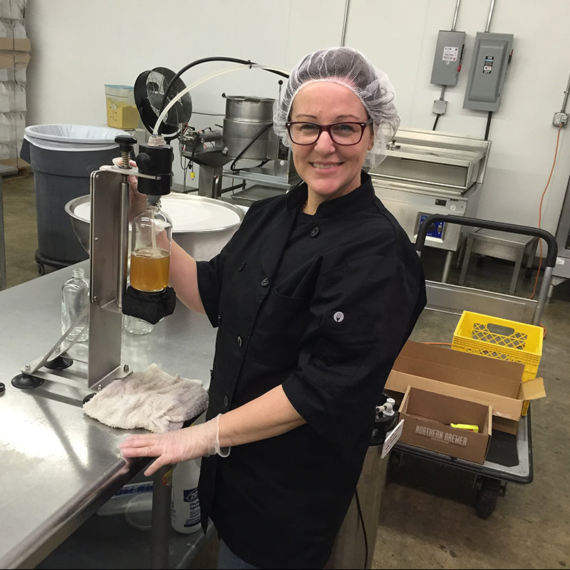 Person in a kitchen or brewery setting, pouring a liquid into a bottle.